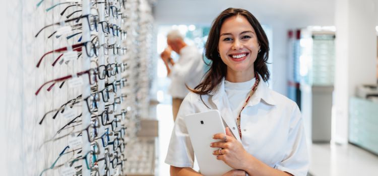 ophthalmology assistant stands next to a display of eyeglasses and smiles at the camera